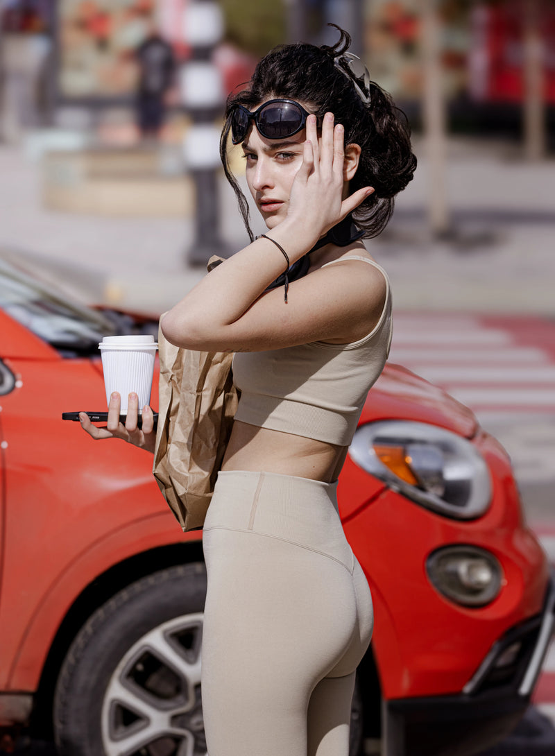 Woman in beige outfit holding a phone and a cup, standing in front of a red car. %Cream% %Hover%